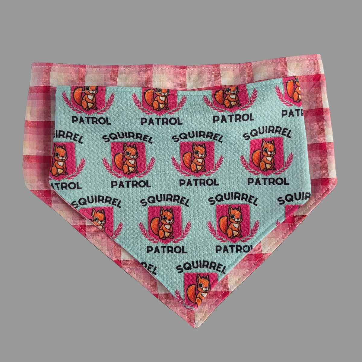 Mint Squirrel Patrol bandana with pink badges laid on its pink plaid reverse side